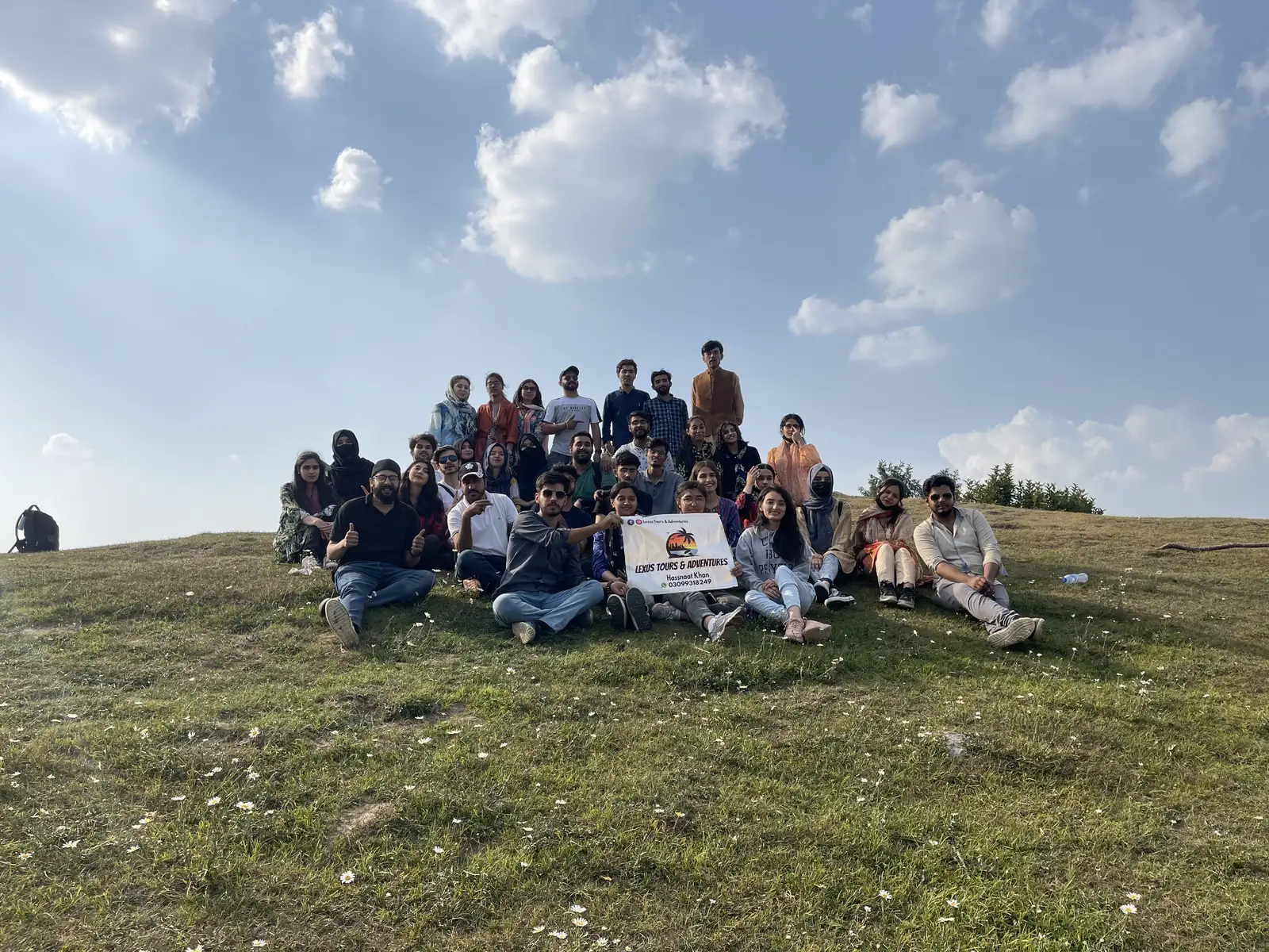 Lexuz Tours group of around thirty travellers sitting on a green meadow holding the company banner